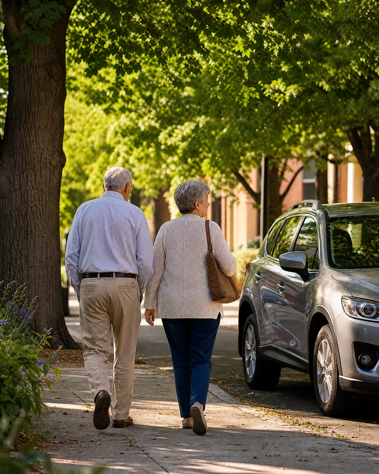 An older man and woman walk away from the camera along a tree-shaded sidewalk beside a parked silver SUV, with a brick building in the background.