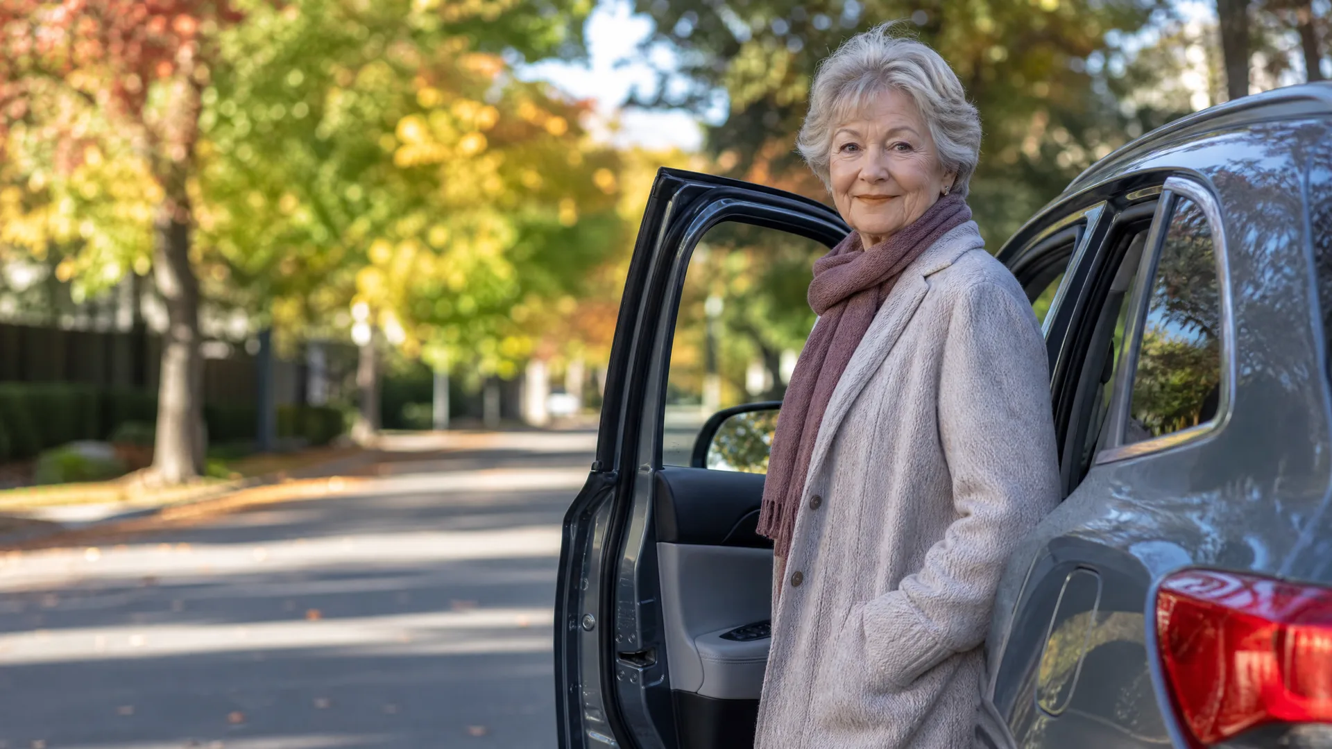 An older woman in a light coat and pink scarf stands beside an open rear door of a dark car on a tree-lined street with autumn foliage.