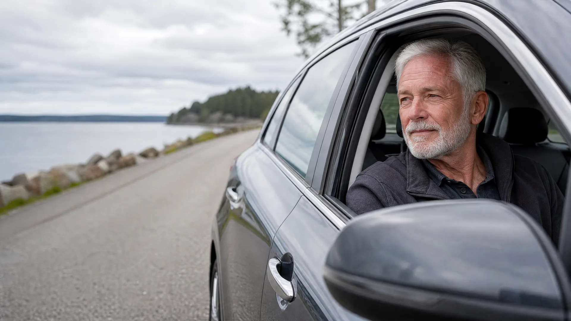An older man with gray hair and a beard sits in the driver's seat of a dark car, looking out the open window, beside a paved road along a rocky shoreline with trees under an overcast sky.
