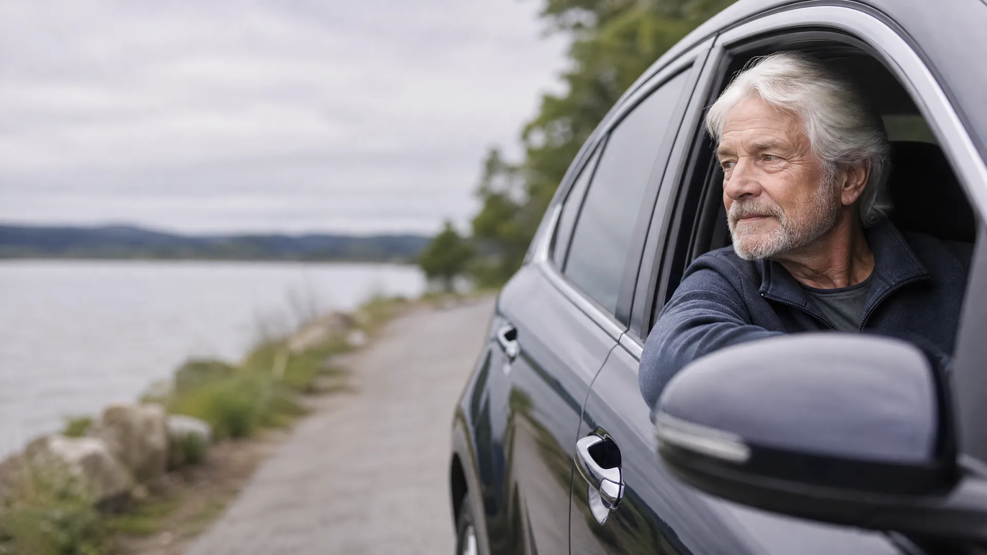 An older man with gray hair and a beard leans his arm out the open driver's window of a dark car parked on a gravel road beside a lake, with low hills and overcast sky in the background.