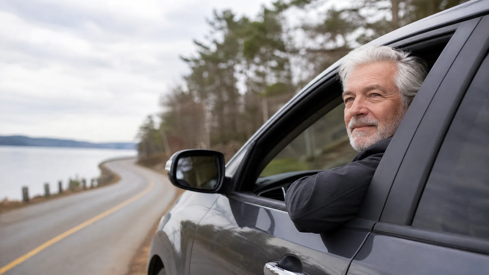 A gray-haired man with a beard leans his arm out of the open window of a dark car parked beside a curving road along a body of water, with conifer trees and an overcast sky behind.