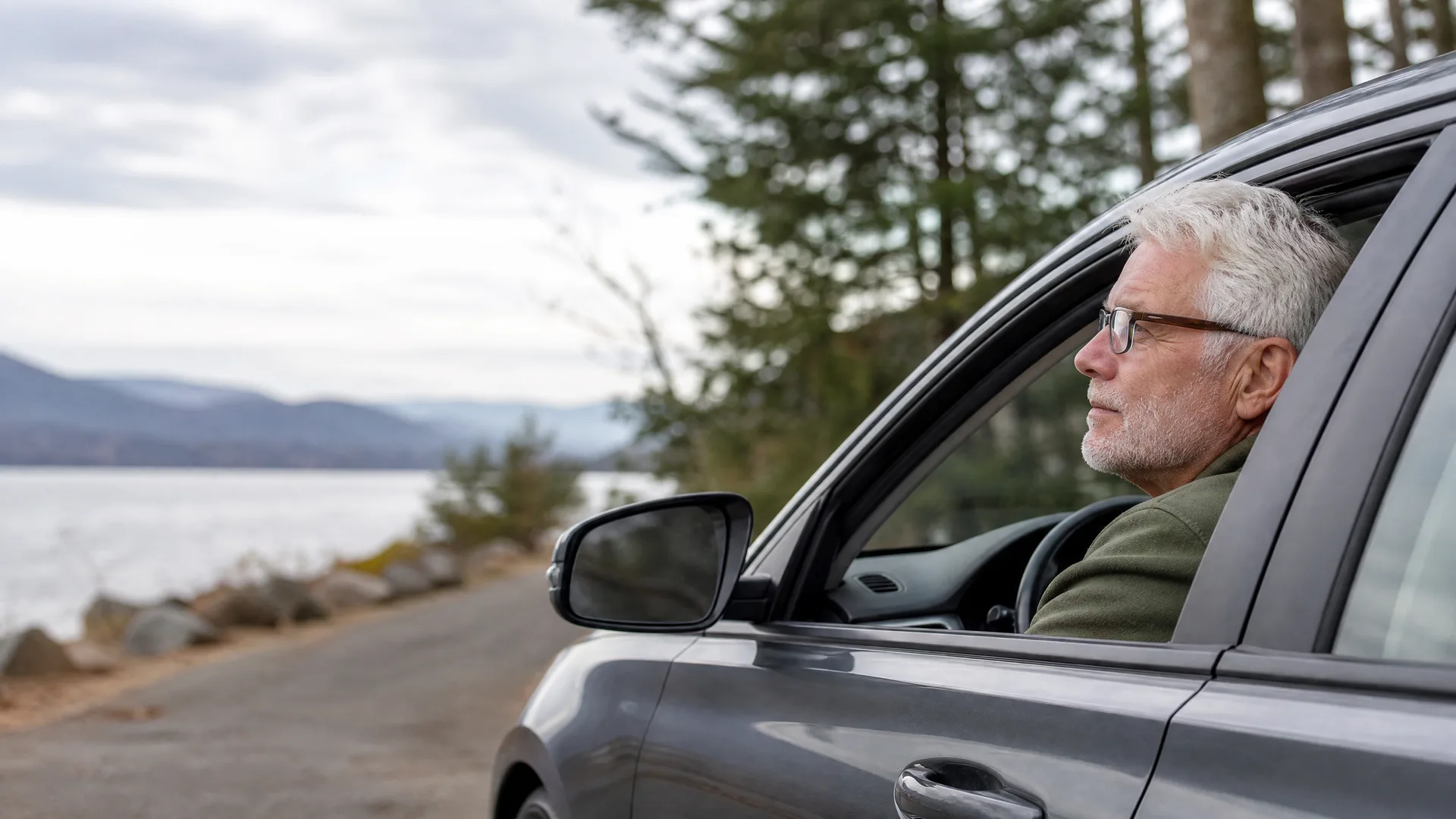 An older man with white hair and glasses sits in the driver's seat of a dark car with the window down, parked on a roadside beside a lake with distant hills and conifer trees under an overcast sky.