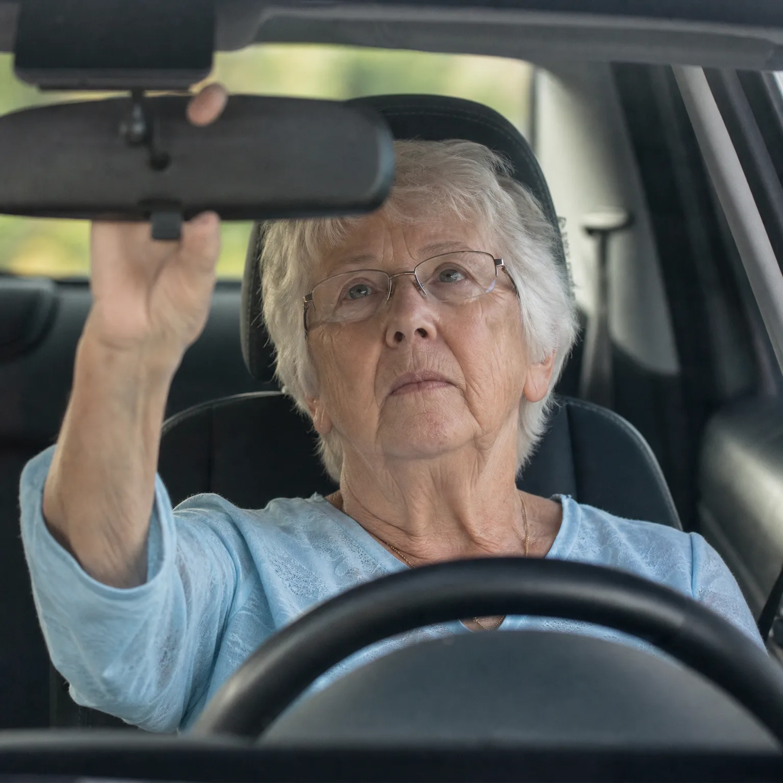 An older woman with short white hair and glasses, wearing a light blue top, sits in a car's driver seat behind the steering wheel and reaches up to adjust the rearview mirror.