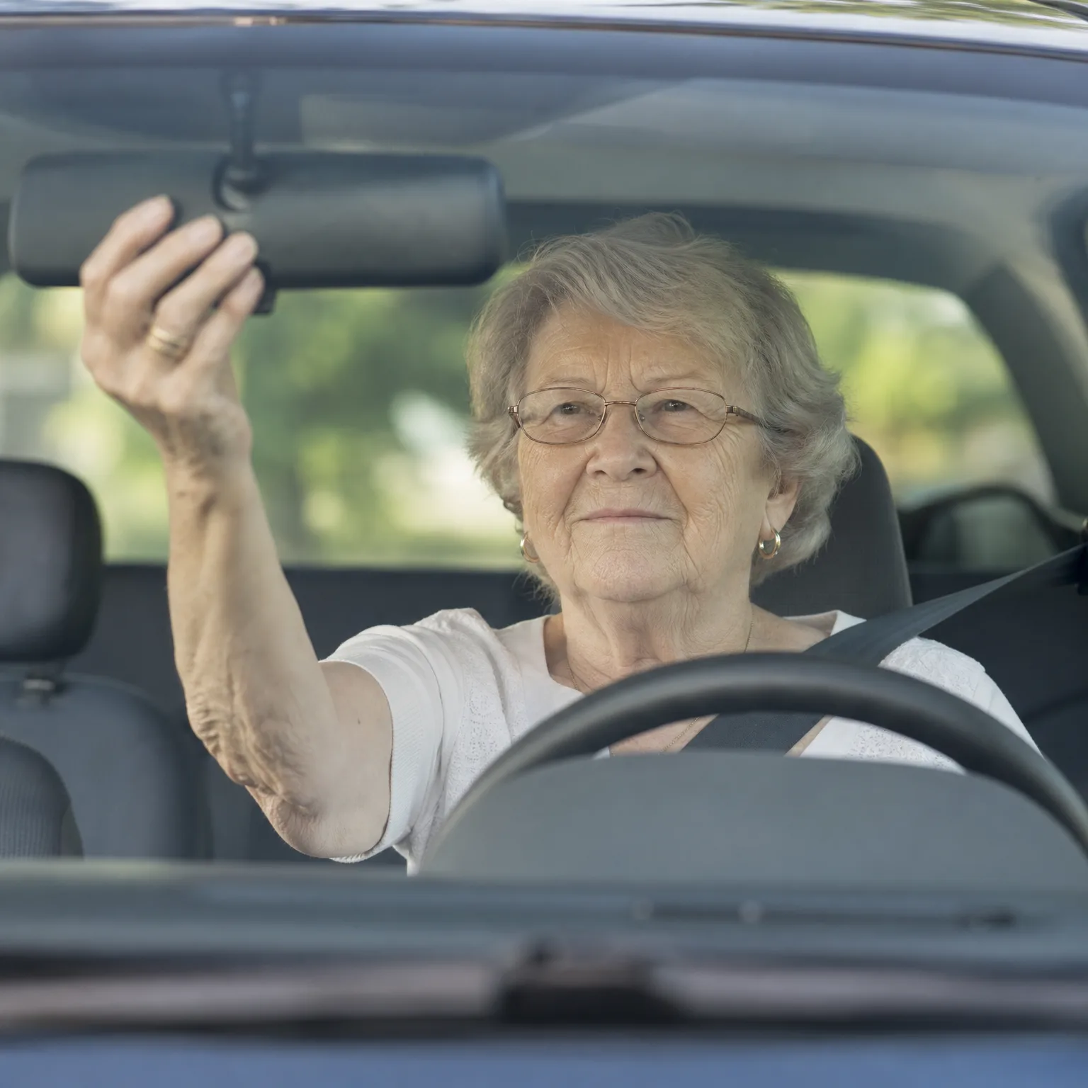 An older woman with glasses sits in a car's driver seat, seen through the windshield, reaching up with her right hand to adjust the rearview mirror.