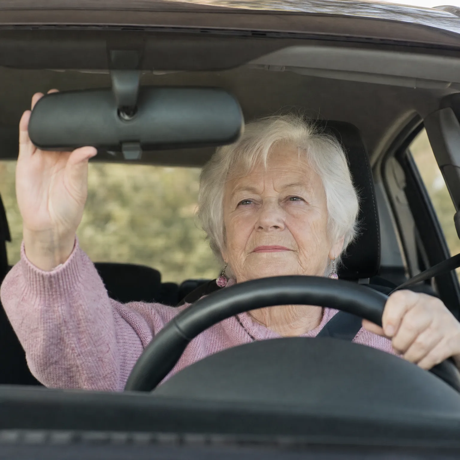 An older woman with short white hair in a pink sweater sits in a car's driver seat, one hand on the steering wheel and the other adjusting the rearview mirror.