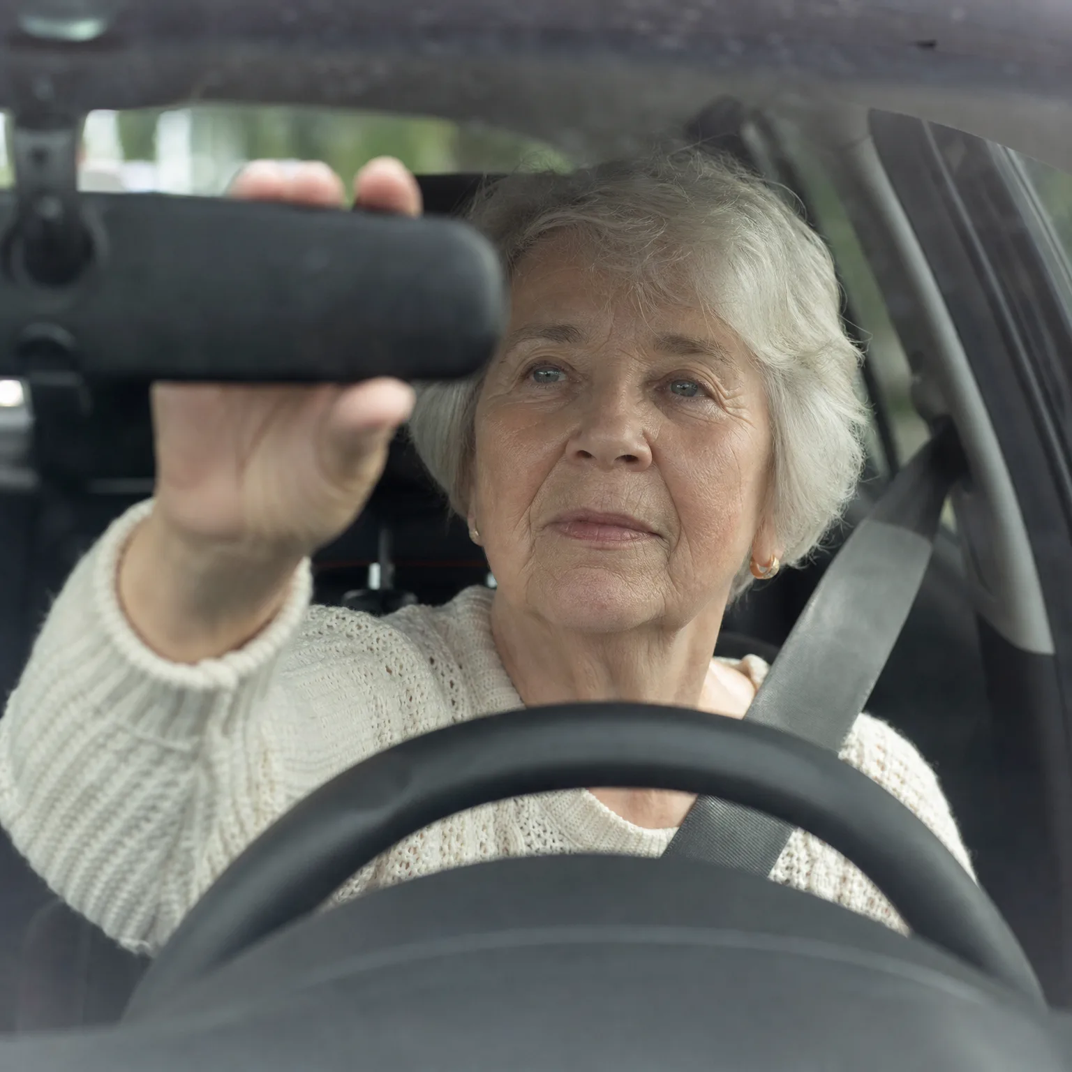An older woman with short grey hair in a cream knit sweater sits in a car's driver seat wearing a seatbelt, reaching up to adjust the rearview mirror, seen through the windshield above the steering wheel.