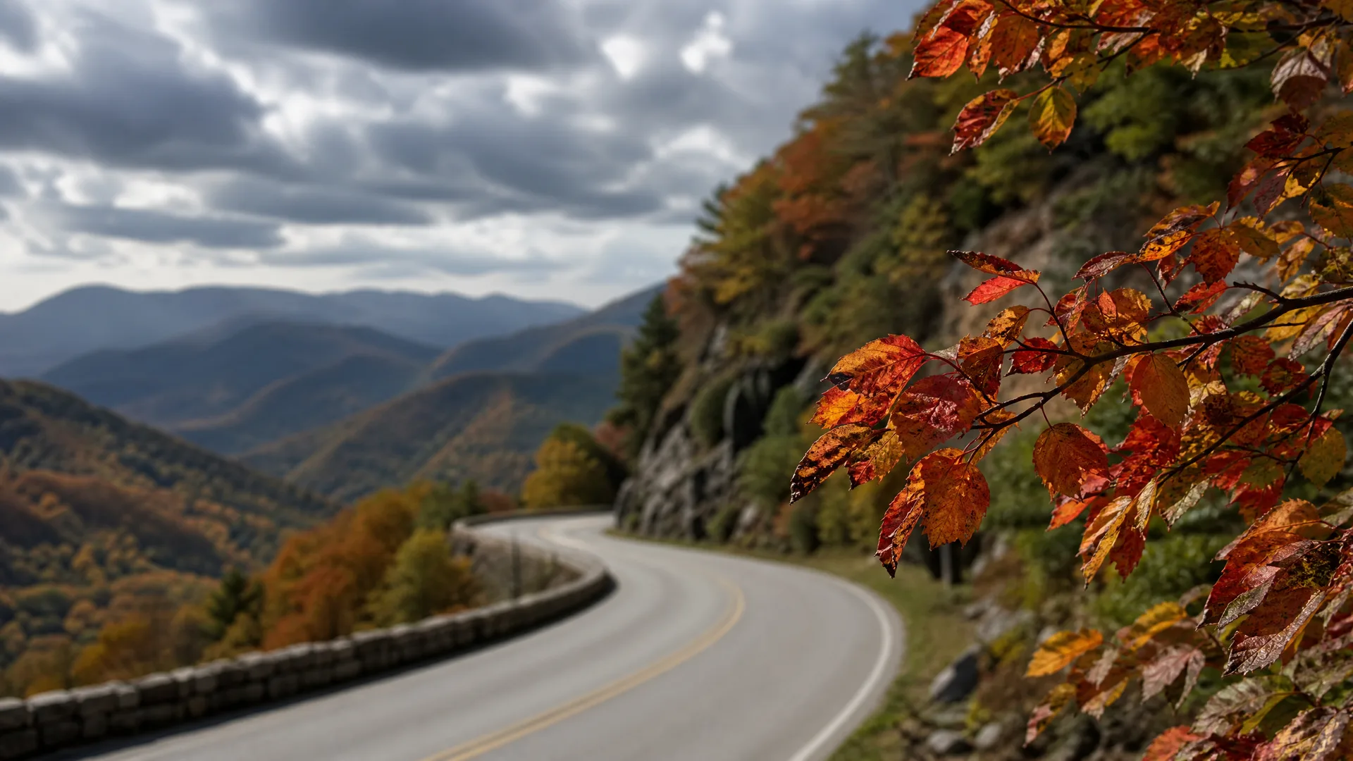 A curving two-lane road with a yellow center line and a low stone wall runs along a hillside, with autumn-colored leaves in the foreground and layered mountain ridges under a cloudy sky in the distance.